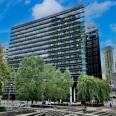 Citysuites Aparthotel Manchester Esterno foto The photo shows a modern multi-story building with a glass and steel facade, surrounded by trees and greenery. The structure features a series of horizontal lines and large windows, reflecting the sky and nearby buildings. In the foreground, there ar