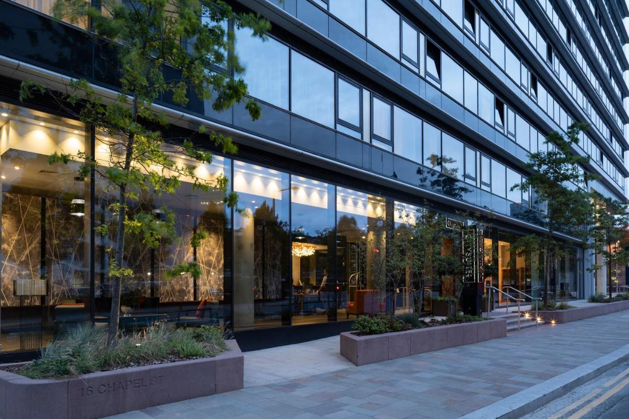 Citysuites Aparthotel Manchester Esterno foto The photo shows a modern building with a glass facade, reflecting the surrounding environment. The structure has several floors and features large windows that allow visibility into the interior, which appears well-lit. There are trees and greenery p