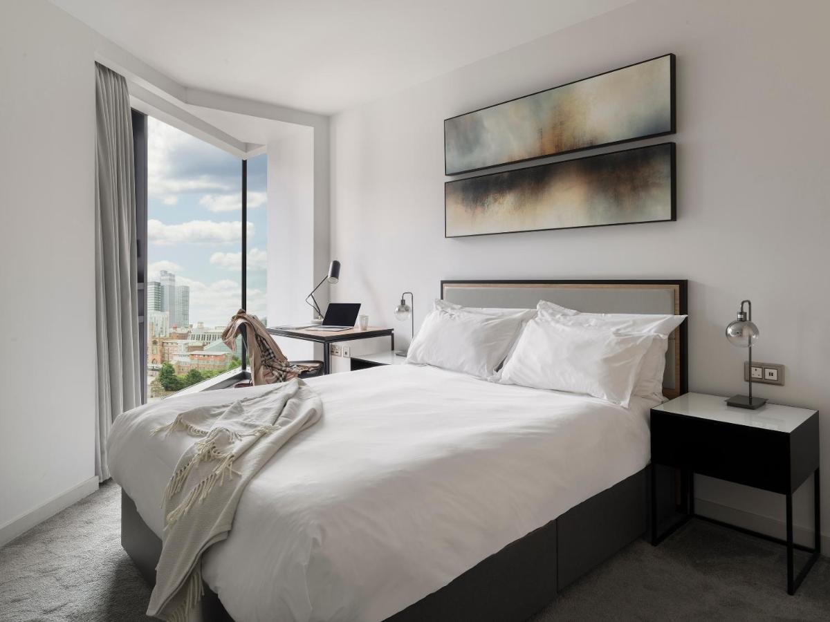 Citysuites Aparthotel Manchester Esterno foto The photo shows a modern bedroom with a minimalistic design. There's a large bed with white bedding, complemented by decorative pillows. To the side of the bed, there are two nightstands, each with a lamp. A stylish piece of art hangs above the bed, 