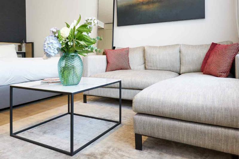 Citysuites Aparthotel Manchester Esterno foto The photo features a modern living area. There is a light-colored, comfortable sectional sofa with decorative cushions in shades of red. A glass coffee table with a black metal frame is positioned in front of the sofa. On the table, there is a striki