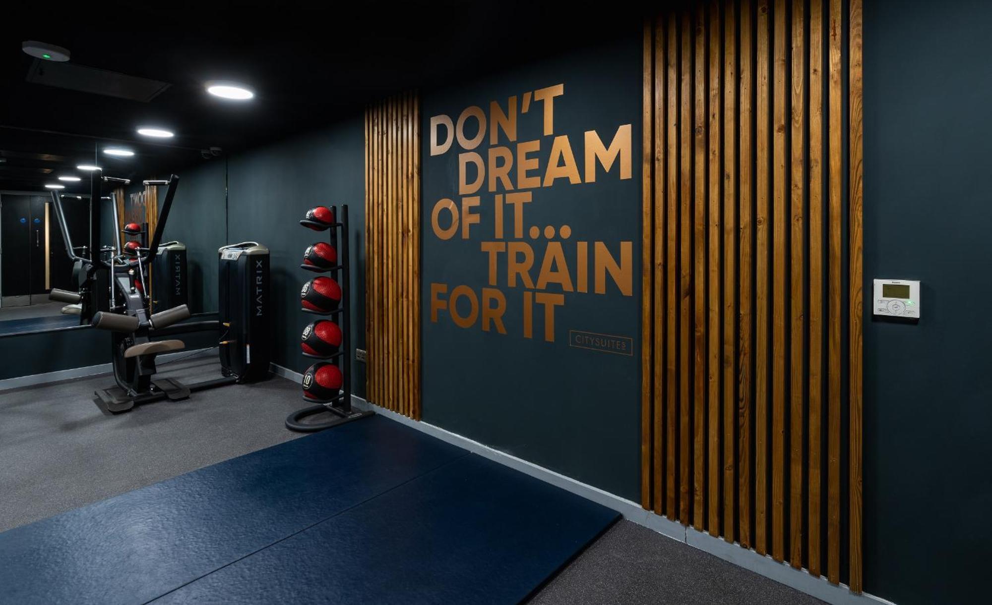 Citysuites Aparthotel Manchester Esterno foto The photo depicts a section of a gym or fitness area. The wall features a motivational quote that reads: "DON'T DREAM OF IT... TRAIN FOR IT," highlighted in large, bold letters. The wall is designed with a combination of dark green paint and wooden s