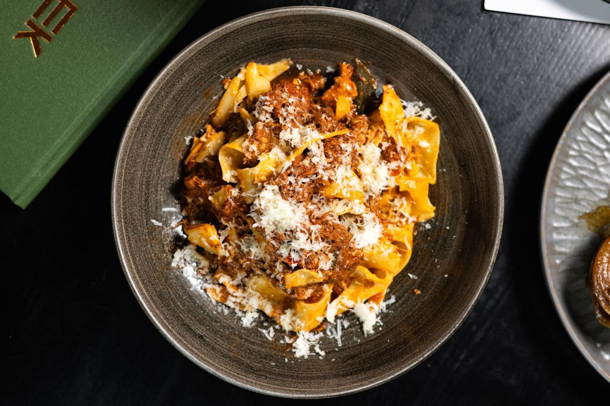Citysuites Aparthotel Manchester Esterno foto The photo features a bowl of pasta, likely pappardelle, topped with a rich meat sauce. The dish is garnished with a generous amount of grated cheese, adding a creamy texture to the meal. It's presented in a dark, round bowl, which highlights the vibr