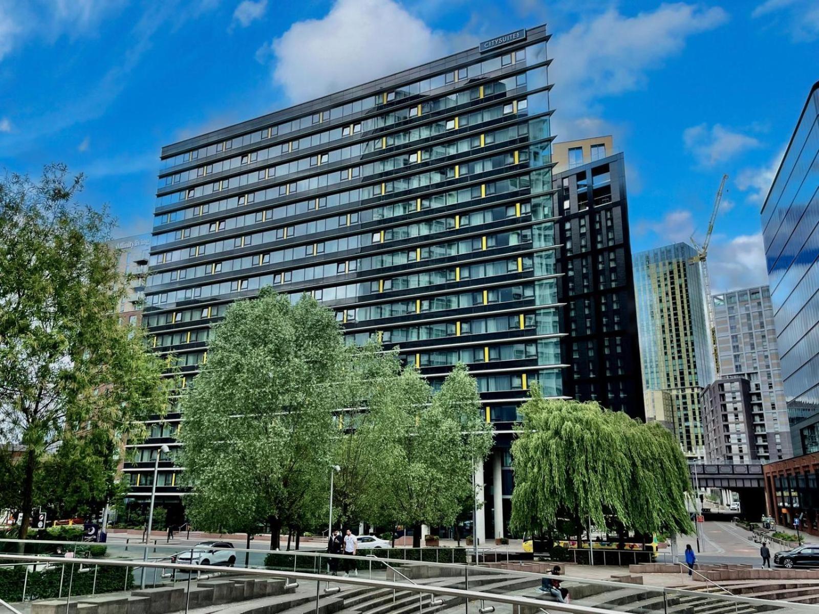 Citysuites Aparthotel Manchester Esterno foto The photo shows a modern multi-story building with a glass and steel facade, surrounded by trees and greenery. The structure features a series of horizontal lines and large windows, reflecting the sky and nearby buildings. In the foreground, there ar