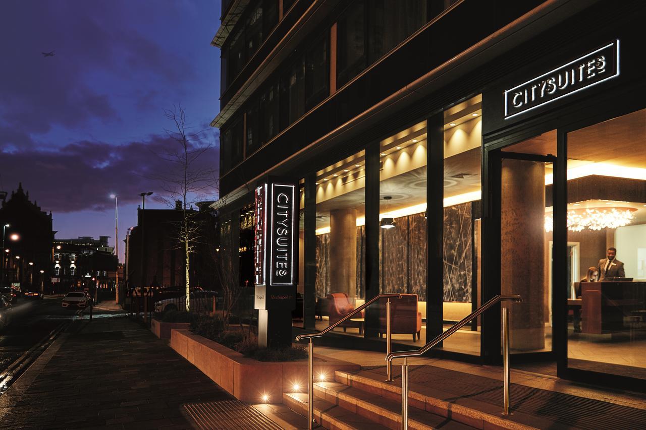 Citysuites Aparthotel Manchester Esterno foto The photo shows the exterior of a modern hotel named "CitySuites," taken at dusk. The entrance features large glass doors and an illuminated sign displaying the hotel's name. The building has a contemporary design with a sleek facade and warm lightin