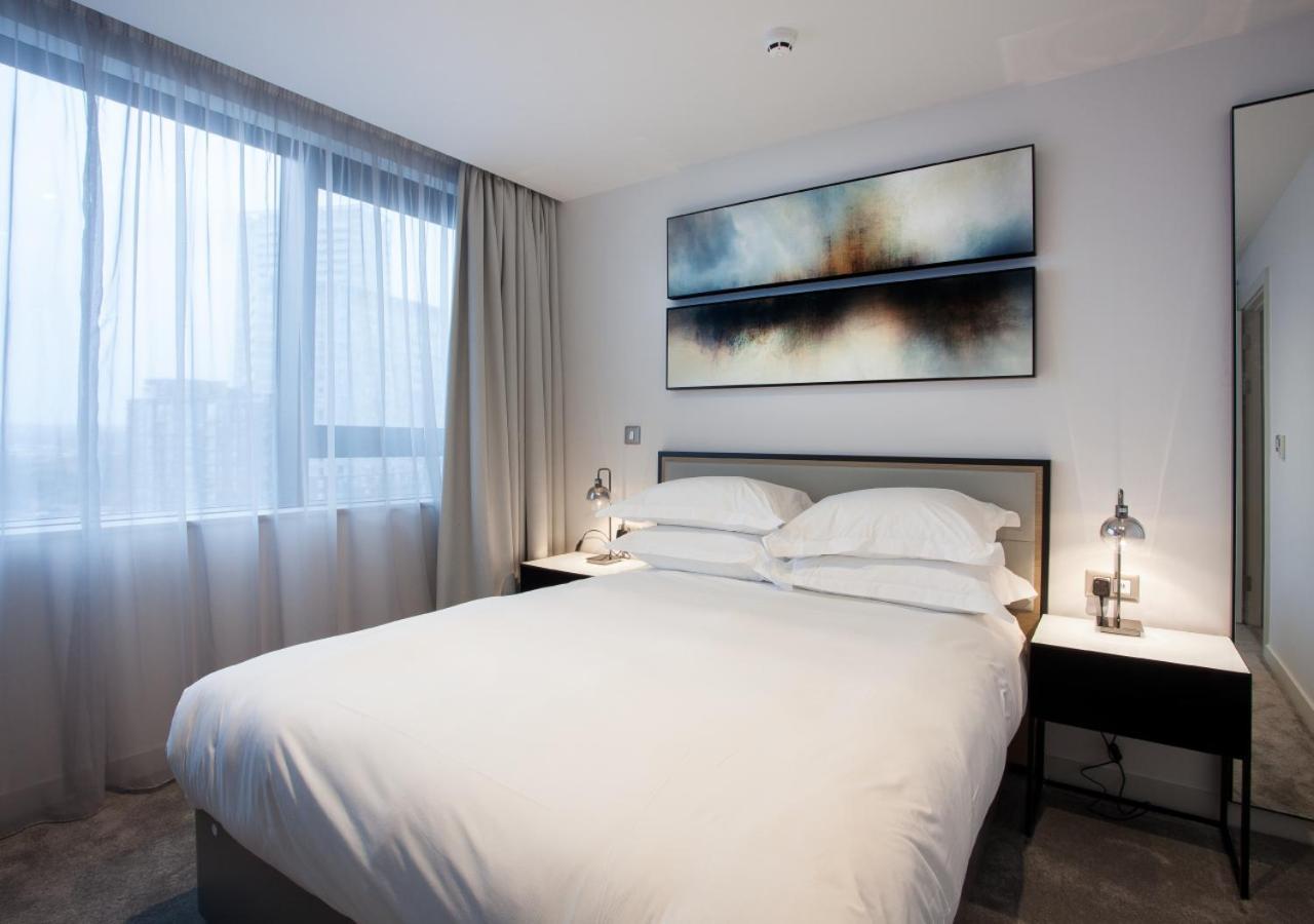 Citysuites Aparthotel Manchester Esterno foto The photo shows a modern bedroom with a double bed featuring white bedding and two pillows. On either side of the bed, there are bedside tables with lamps. Above the bed, there is a piece of abstract artwork. The room has large windows covered with s