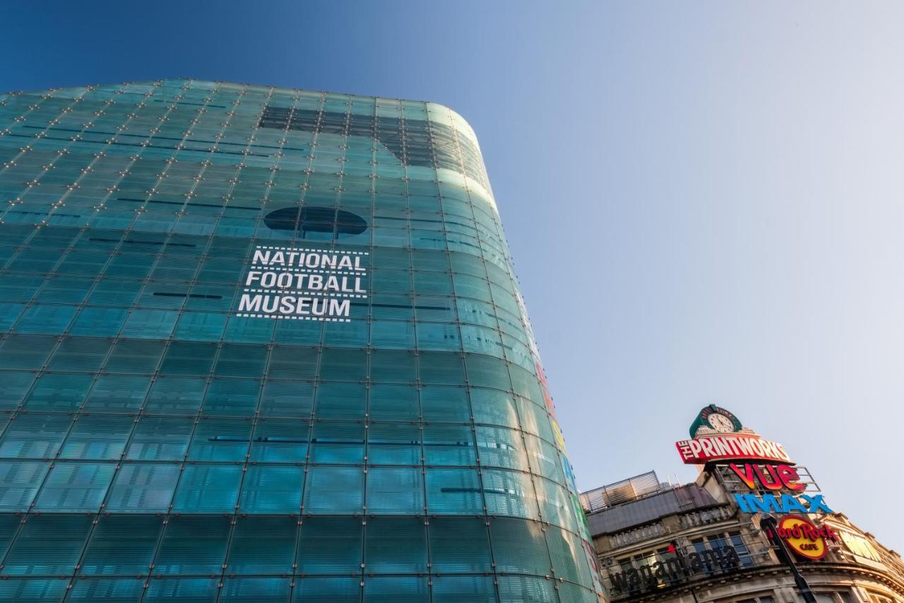 Citysuites Aparthotel Manchester Esterno foto The photo shows a modern glass building with the words "NATIONAL FOOTBALL MUSEUM" prominently displayed on one side. The building has a sleek, contemporary design, reflecting light in various shades. In the foreground, there is a colorful sign on a n