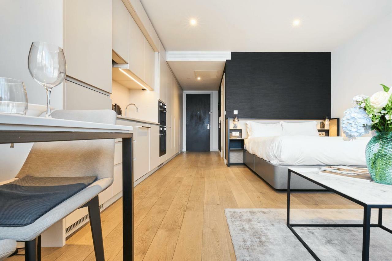 Citysuites Aparthotel Manchester Esterno foto The photo shows a modern studio apartment. The space features a compact kitchen area with cabinetry and appliances on one side, and a neatly made bed against a dark accent wall. There is a light wooden floor that adds warmth to the room. A small tabl