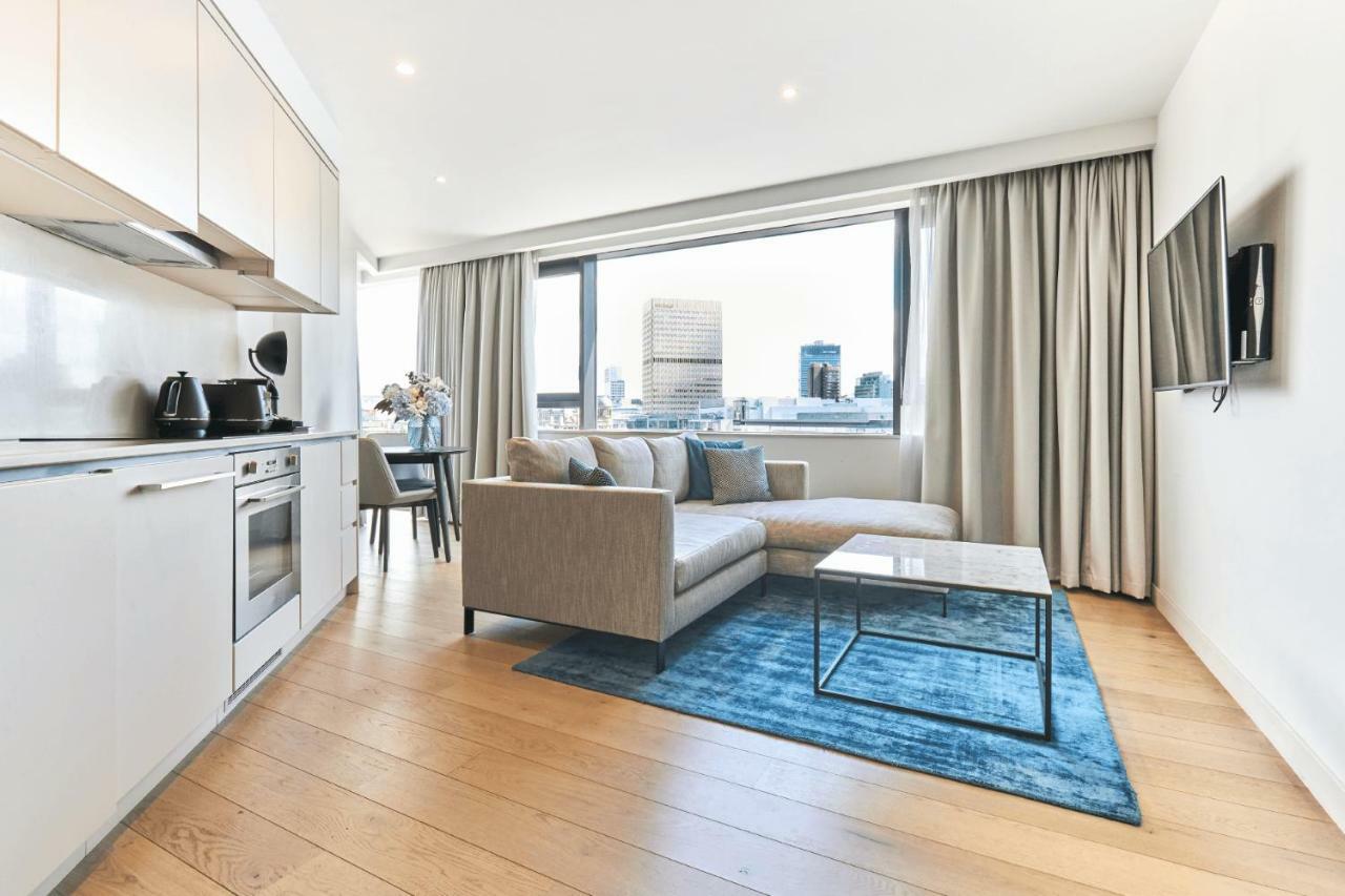 Citysuites Aparthotel Manchester Esterno foto The photo shows a modern living space with a stylish design. On one side, there is a kitchenette featuring a stove, oven, kettle, and other appliances, all in a light color. The main living area includes a light gray sofa positioned on a blue area ru
