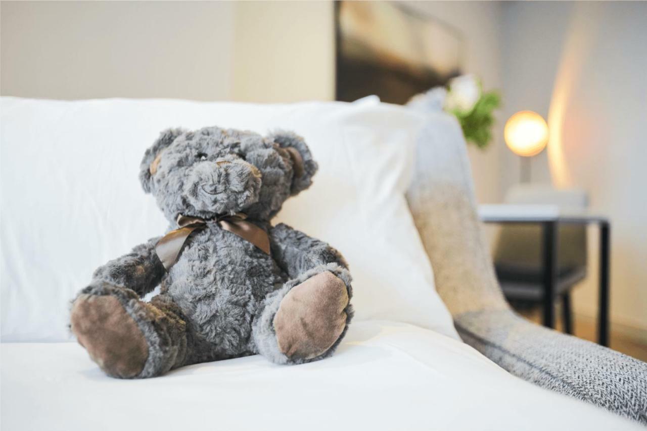 Citysuites Aparthotel Manchester Esterno foto The photo shows a plush teddy bear, which is gray in color, sitting on a white couch. The bear has a brown ribbon tied around its neck and has seen some wear, with noticeable shading on its paws. In the background, there is a softly lit environment w